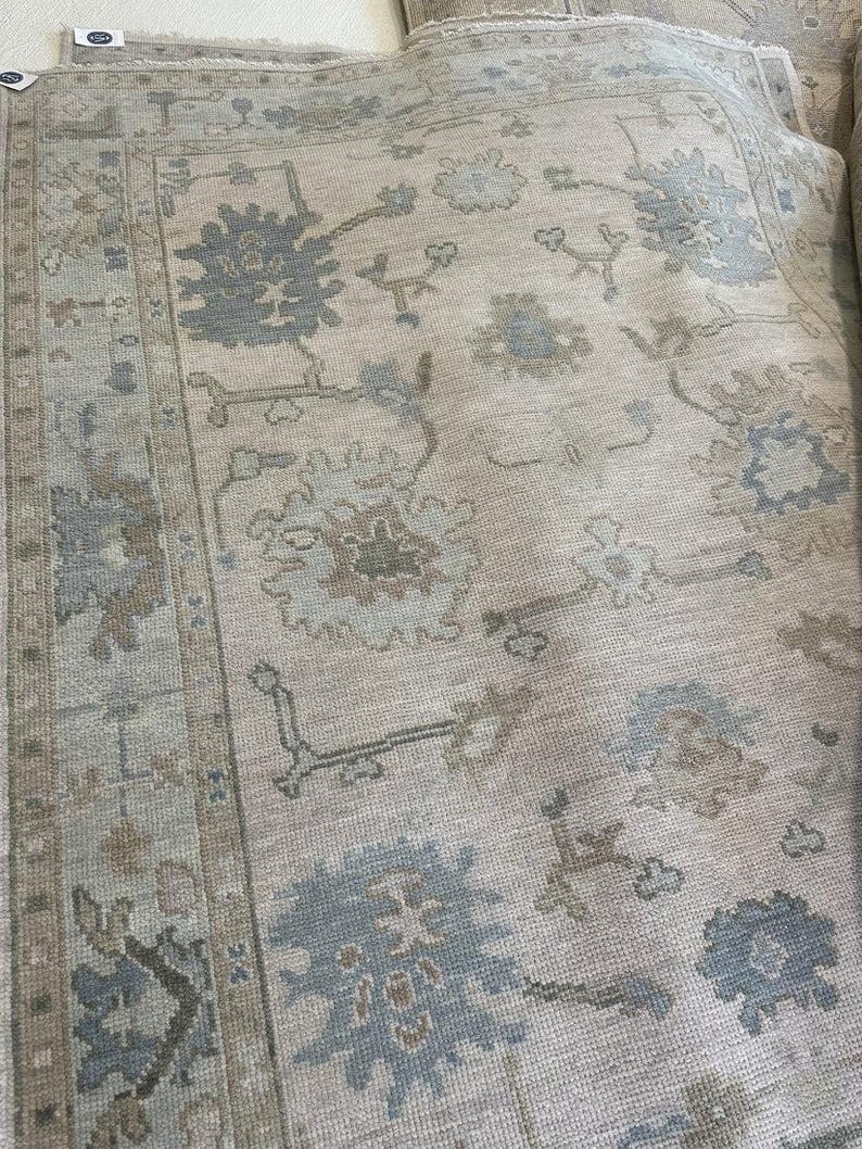 K&ouml;nnte beinhalten: Ein gro&szlig;er Teppich mit hellbeigem Hintergrund und einem wiederkehrenden Muster aus Blumen- und Rankenmotiven in gedeckten Schattierungen von Schieferblau, Taupe und sanftem Grau. Der Teppich hat eine breite Bord&uuml;re mit geometrischen Elementen.