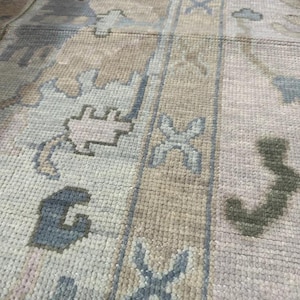 K&ouml;nnte beinhalten: Nahaufnahme eines rechteckigen Teppichs mit geometrischem Muster. Der Teppich weist eine Kombination aus hellblauen, beigen und olivgr&uuml;nen Farben auf. Das Design umfasst stilisierte Blumen- und geometrische Formen und erzeugt eine strukturierte Oberfl&auml;che.