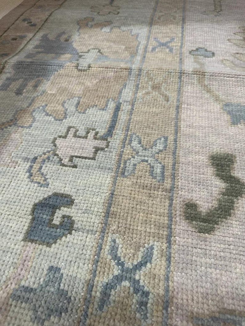 K&ouml;nnte beinhalten: Nahaufnahme eines rechteckigen Teppichs mit geometrischem Muster. Der Teppich weist eine Kombination aus hellblauen, beigen und olivgr&uuml;nen Farben auf. Das Design umfasst stilisierte Blumen- und geometrische Formen und erzeugt eine strukturierte Oberfl&auml;che.
