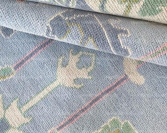 May include: Close-up of a rolled-up, patterned rug. The rug features a light blue background with a repeating floral design in shades of peach, green, and white. Pink and white lines accent the design, creating a textured appearance.