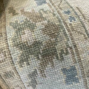 K&ouml;nnte beinhalten: Nahaufnahme eines Abschnitts eines gewebten Teppichs mit detailliertem Muster. Der Teppich weist eine Mischung aus neutralen Farben auf, darunter Beige, Grau und ged&auml;mpftes Blau, wodurch ein weiches, strukturiertes Aussehen entsteht. Das Design integriert geometrische und florale Elemente.