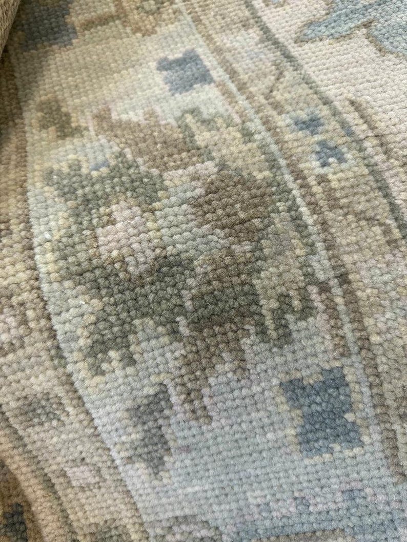 K&ouml;nnte beinhalten: Nahaufnahme eines Abschnitts eines gewebten Teppichs mit detailliertem Muster. Der Teppich weist eine Mischung aus neutralen Farben auf, darunter Beige, Grau und ged&auml;mpftes Blau, wodurch ein weiches, strukturiertes Aussehen entsteht. Das Design integriert geometrische und florale Elemente.