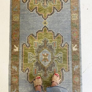 May include: A rectangular rug with a central design in shades of green, brown, and orange, set against a light blue background. The rug has a border in shades of brown and orange. The rug is on a white surface.