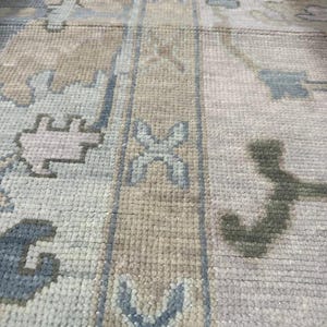 K&ouml;nnte beinhalten: Nahaufnahme eines beige-grauen Teppichs mit Muster. Der Teppich weist ein sich wiederholendes geometrisches Design mit blauen und hellbraunen Akzenten auf. Die Textur des Teppichs ist sichtbar, was auf eine gewebte Konstruktion hindeutet. Die Gesamt&auml;sthetik ist neutral und f&uuml;r verschiedene Einrichtungsstile geeignet.