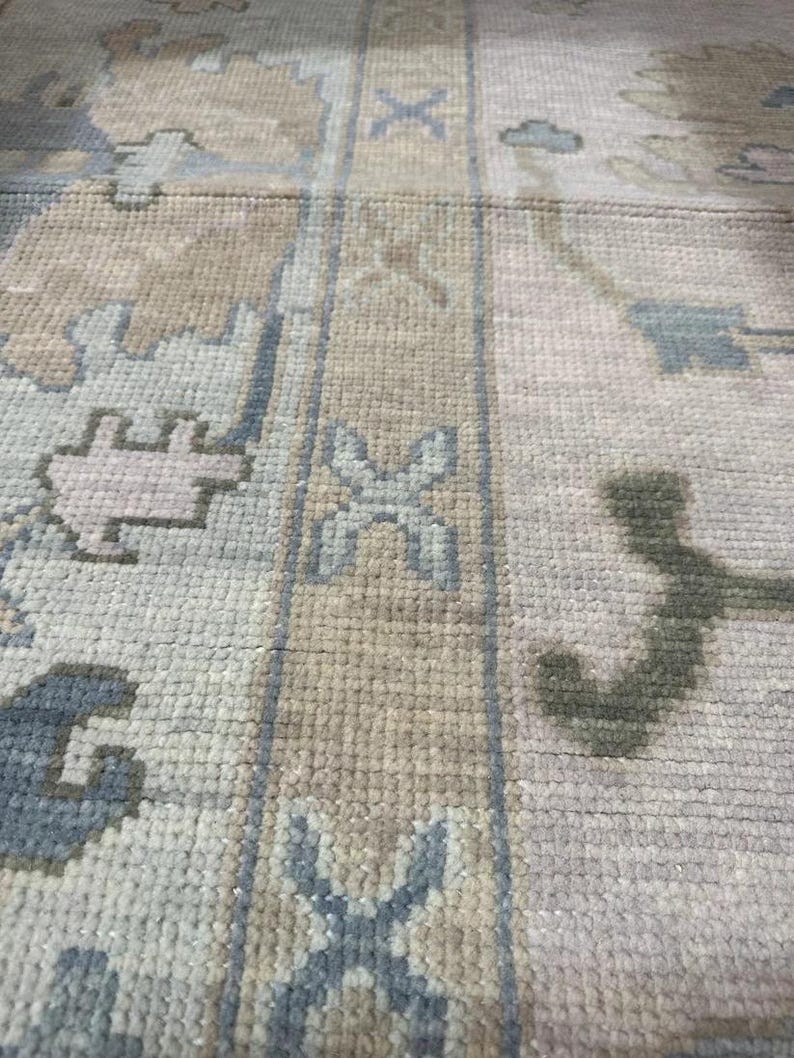 K&ouml;nnte beinhalten: Nahaufnahme eines beige-grauen Teppichs mit Muster. Der Teppich weist ein sich wiederholendes geometrisches Design mit blauen und hellbraunen Akzenten auf. Die Textur des Teppichs ist sichtbar, was auf eine gewebte Konstruktion hindeutet. Die Gesamt&auml;sthetik ist neutral und f&uuml;r verschiedene Einrichtungsstile geeignet.
