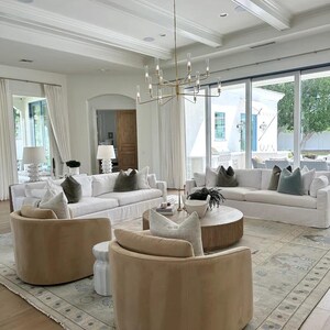 K&ouml;nnte beinhalten: Ein ger&auml;umiges Wohnzimmer mit wei&szlig;en Sofas, beigen Drehst&uuml;hlen und einem runden Couchtisch aus Holz. Der Raum verf&uuml;gt &uuml;ber einen gro&szlig;en Teppich, einen goldenen Kronleuchter und gro&szlig;e Fenster.
