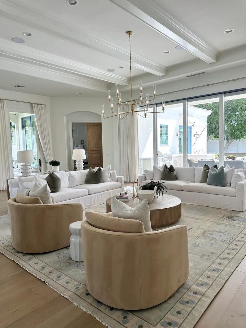 K&ouml;nnte beinhalten: Ein ger&auml;umiges Wohnzimmer mit wei&szlig;en Sofas, beigen Drehst&uuml;hlen und einem runden Couchtisch aus Holz. Der Raum verf&uuml;gt &uuml;ber einen gro&szlig;en Teppich, einen goldenen Kronleuchter und gro&szlig;e Fenster.