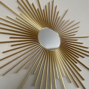 May include: A gold-colored sunburst mirror with a circular mirror in the center. The sunburst design features numerous straight, radiating lines. The mirror is mounted on a textured, off-white wall.