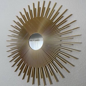 May include: A gold-colored sunburst mirror with a circular mirror in the center. The sunburst design features numerous straight, radiating rods of varying lengths. The mirror is mounted on a light gray wall.