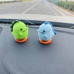 May include: Two crocheted dinosaur figurines, one green and one blue, each with a small wooden base. The green dinosaur has a green spiky back and the blue dinosaur has a blue spiky back. Both have black eyes and smiling faces.