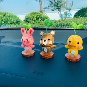 May include: Three crocheted animal figurines: a pink bunny, a brown bear, and a yellow duck. Each figure has a green sprout on its head and sits on a small wooden base. The bunny and duck have pink cheeks.