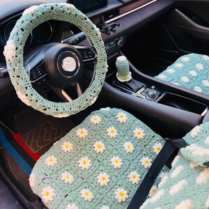 May include: A crochet car interior set in a light green color, featuring white and yellow daisy accents. The set includes a steering wheel cover, gear shift cover, and seat covers. The crochet work is detailed and adds a unique, handmade touch to the car's interior.