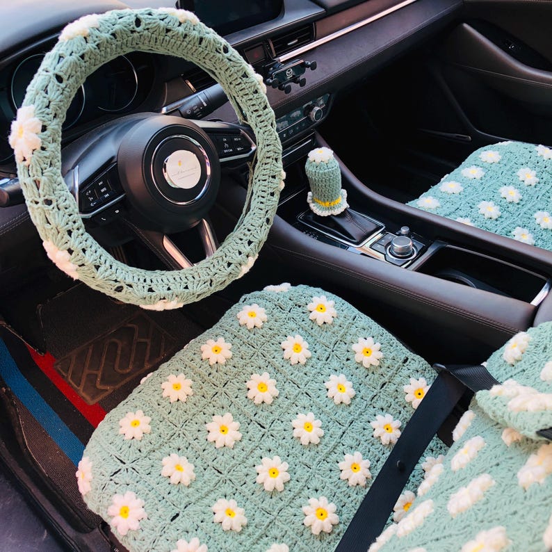 May include: A crochet car interior set in a light green color, featuring white and yellow daisy accents. The set includes a steering wheel cover, gear shift cover, and seat covers. The crochet work is detailed and adds a unique, handmade touch to the car's interior.