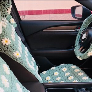 May include: Crocheted car seat covers and steering wheel cover in a light green color, adorned with white and yellow daisy flower appliques. The interior of a car is visible, with the seat and steering wheel cover in focus.