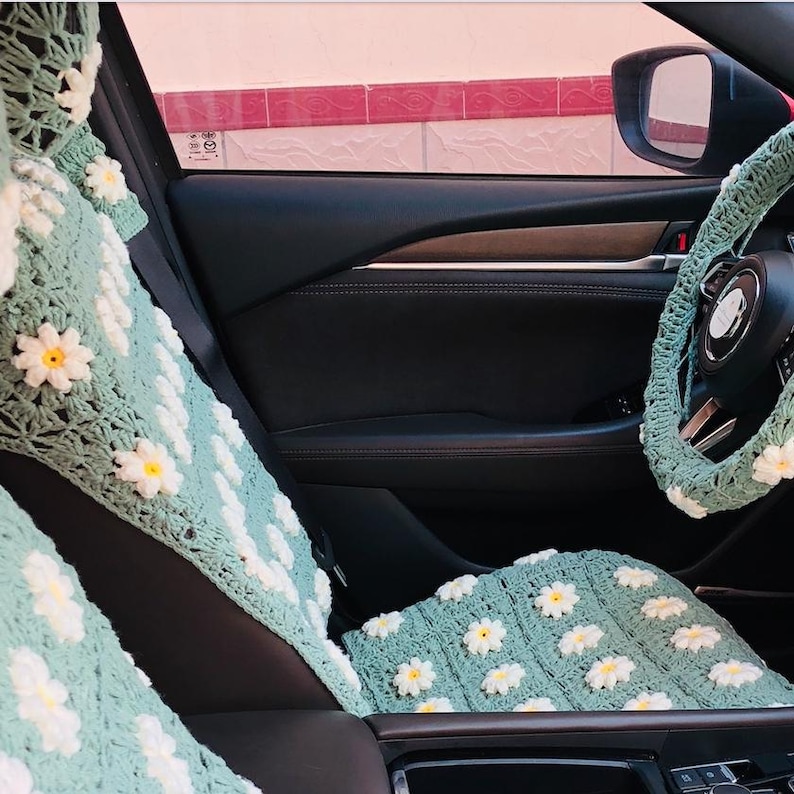 May include: Crocheted car seat covers and steering wheel cover in a light green color, adorned with white and yellow daisy flower appliques. The interior of a car is visible, with the seat and steering wheel cover in focus.