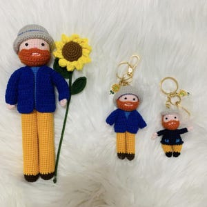 Könnte beinhalten: Gehäkelte Puppe und Schlüsselanhänger, die einem Mann mit rotem Bart, blauer Jacke und gelber Hose ähneln. Eine gehäkelte Sonnenblume ist ebenfalls vorhanden. Die Puppe ist etwa 30 cm groß.