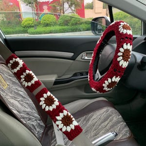 Könnte beinhalten: Gehäkeltes Auto-Zubehör-Set, bestehend aus einem roten Lenkradbezug und einem Sicherheitsgurtbezug. Beide sind mit weißen und braunen Blumenmustern verziert. Der Innenraum des Autos ist im Hintergrund zu sehen.