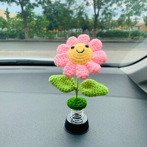 May include: A handmade crochet flower with a smiling yellow center and pink petals. The flower has green leaves and a green base, mounted on a black spring base. The flower is a decorative item.
