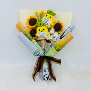 May include: A handmade bouquet featuring crocheted sunflowers, yellow pineapple characters with green tops, and white flowers. The bouquet is wrapped in newspaper-style paper and tied with a brown ribbon. The pineapple characters have the text "Fine Apple It's fine. You're fine. Everything's fine."