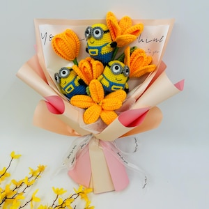 Może przedstawiać: Bukiet szydełkowych kwiatów z trzema postaciami Minionków, żółtymi tulipanami i żółtym kwiatem. Bukiet jest owinięty w papier w kolorze brzoskwiniowym i kremowym i zawiązany wstążką. Na papierze widoczny jest napis "You shine with you".