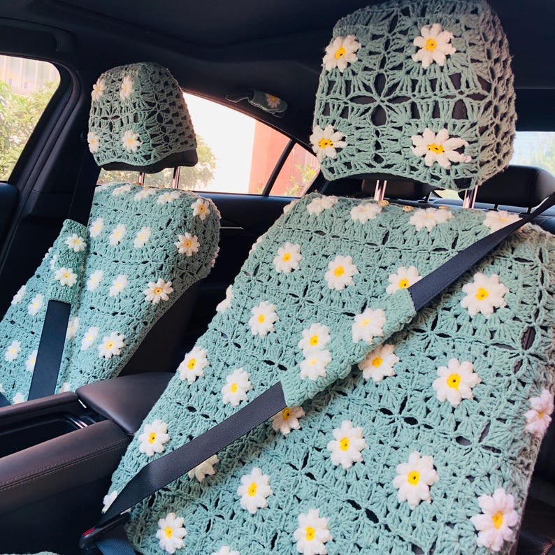 May include: Crocheted car seat covers in a light green color, adorned with white and yellow daisy flowers. The set includes covers for the headrests and seat cushions, with a visible seat belt. The crochet pattern is open and airy.