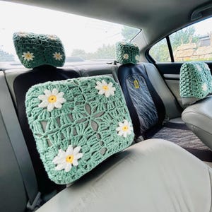 May include: Crocheted car seat covers in a light green color, adorned with white and yellow daisy accents. The set includes headrest covers and a square seat cushion, offering a handmade, floral aesthetic for vehicle interiors.