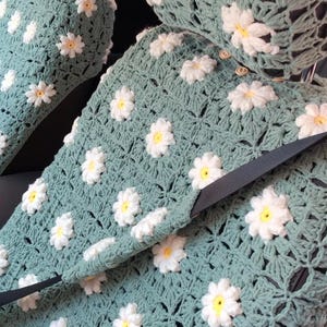 May include: Crocheted car seat covers in a soft teal color, adorned with white and yellow daisy appliques. The covers include headrest covers and seat belt access, offering a handmade, floral design for vehicle interiors.
