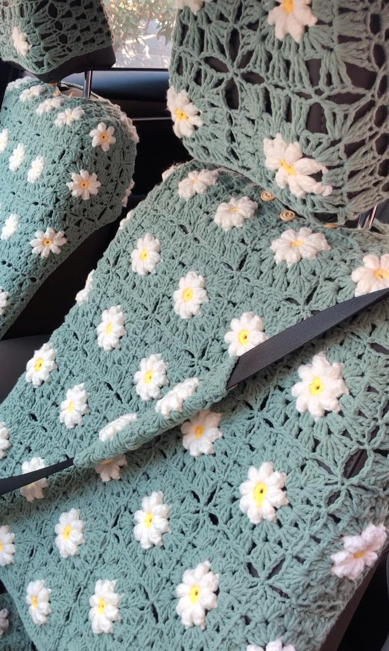 May include: Crocheted car seat covers in a soft teal color, adorned with white and yellow daisy appliques. The covers include headrest covers and seat belt access, offering a handmade, floral design for vehicle interiors.