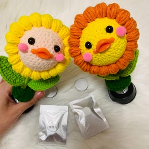 May include: Two handmade crochet sunflower-duck hybrids. One has yellow petals, the other orange. Both have pink cheeks, black eyes, and orange beaks. Green leaves and stems support the flowers. Two small silver packets and two white circles are also present.