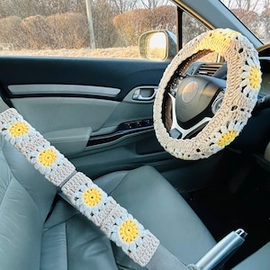 Puede incluir: Juego de accesorios de coche de ganchillo con una funda para el volante y una funda para el cinturón de seguridad. Las fundas son de color beige con diseños de margaritas blancas y amarillas. Los accesorios están en un coche.