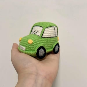 May include: A handmade, green crocheted car toy with a white roof, black wheels, and yellow headlights. The toy car is held in a hand, showcasing its compact size and detailed craftsmanship. The car's windows are white, and the body is a vibrant green.