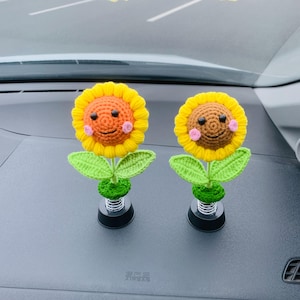 May include: Two crocheted sunflower decorations with smiling faces, yellow petals, and orange centers. The sunflowers have green leaves and sit on black bases with springs. These are likely car accessories.