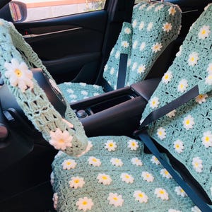 May include: Crocheted car seat covers and steering wheel cover in a light green color, adorned with white and yellow daisy flowers. The set includes covers for the front seats and headrests, creating a coordinated floral theme.