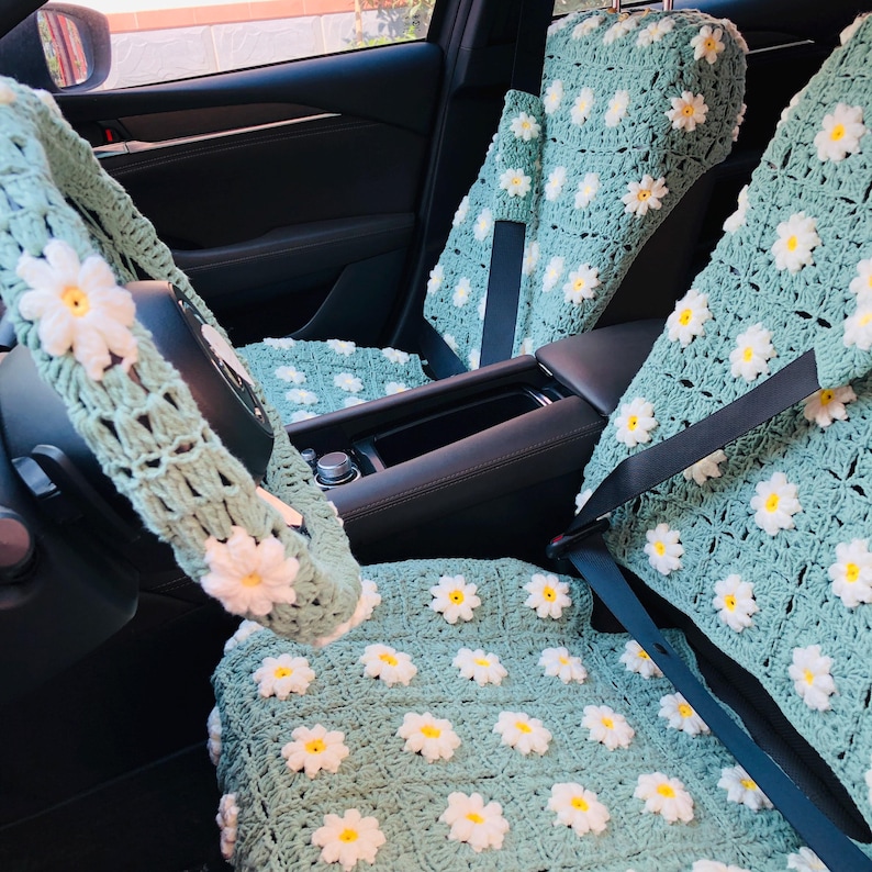 May include: Crocheted car seat covers and steering wheel cover in a light green color, adorned with white and yellow daisy flowers. The set includes covers for the front seats and headrests, creating a coordinated floral theme.