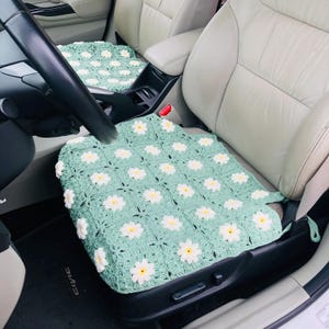 May include: A crocheted car seat cover in a daisy pattern. The cover is light green with white and yellow daisy flowers. The cover is placed on a car seat.