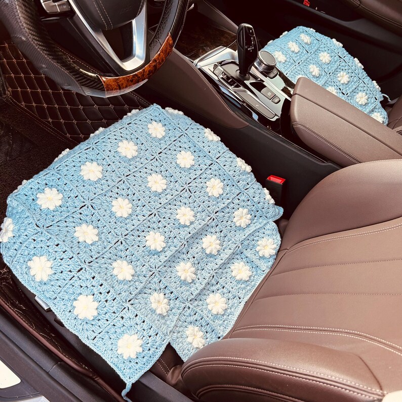 May include: Light blue crochet car seat cover with white flower accents. The cover is placed on a brown leather car seat. The steering wheel and dashboard are visible in the background. The crochet pattern is square.