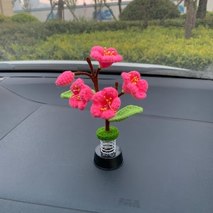 May include: A handcrafted crochet flower arrangement with bright pink flowers, yellow centres, and green leaves. The flowers are attached to a brown stem and sit on a green base with a silver spring and black base. This decorative item is likely for a car dashboard.