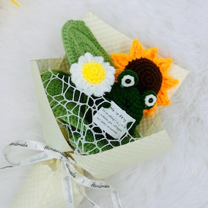 May include: A handmade crochet bouquet featuring a green frog, a sunflower, and a daisy. The frog has large eyes and a tag that reads "He to Fris". The bouquet is wrapped in cream-colored paper and tied with a ribbon that says "handmade".