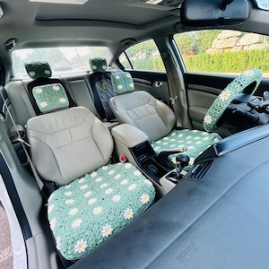 May include: A car interior featuring light green crochet seat covers and steering wheel cover, adorned with white and yellow daisy patterns. The set includes covers for the front and back seats, headrests, and steering wheel, creating a coordinated, handmade aesthetic.