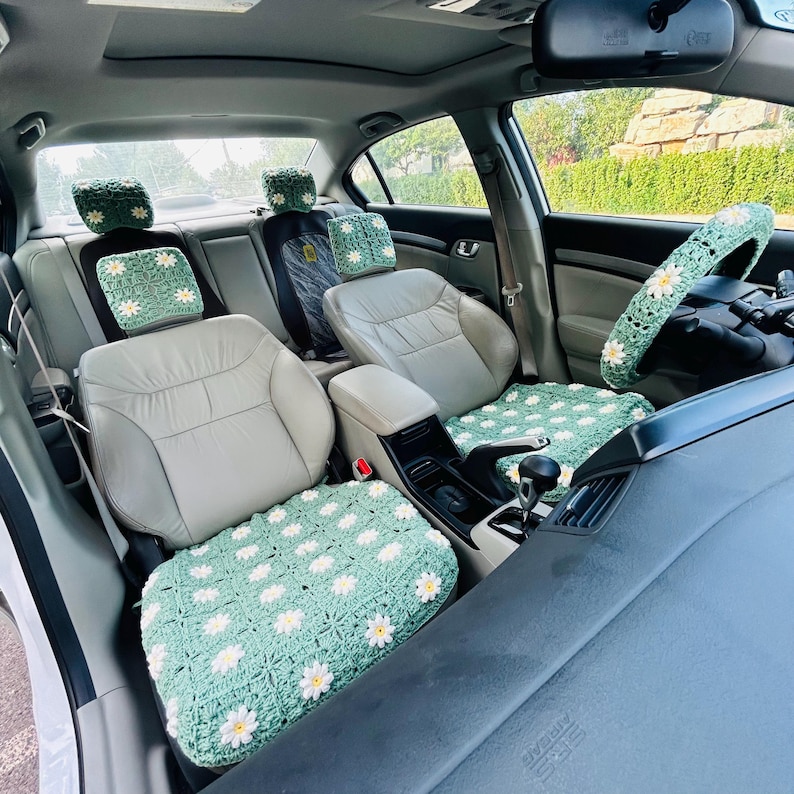 May include: A car interior featuring light green crochet seat covers and steering wheel cover, adorned with white and yellow daisy patterns. The set includes covers for the front and back seats, headrests, and steering wheel, creating a coordinated, handmade aesthetic.