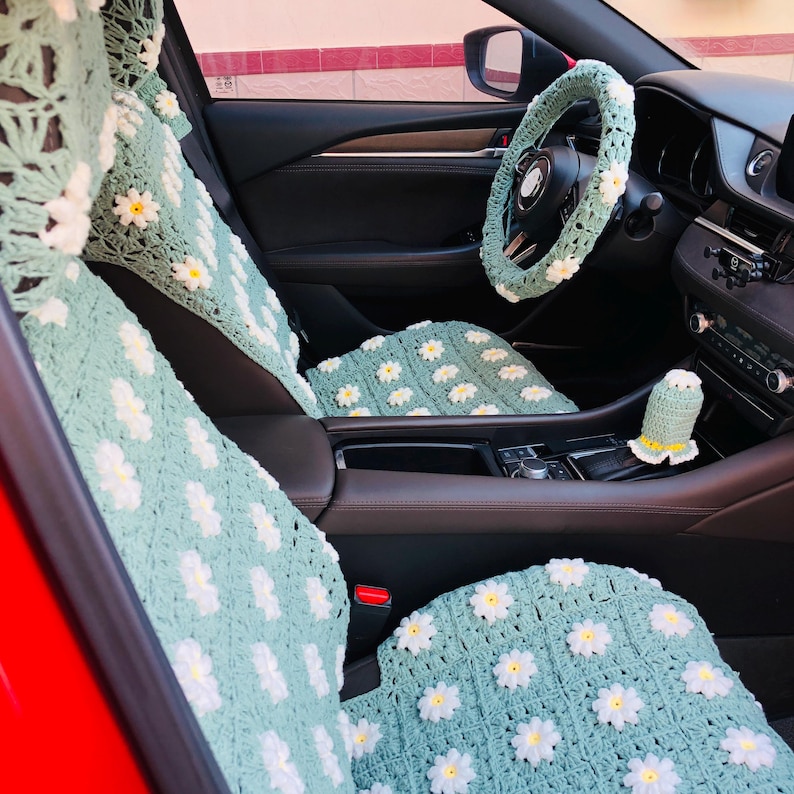May include: Crocheted car seat covers, steering wheel cover, and gear shift cover in a light green color with white and yellow daisy accents. The interior of the car is visible, with a red exterior.