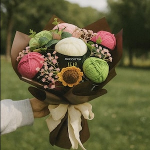 May include: A bouquet of yarn balls in shades of pink, green, and white, wrapped in brown paper and tied with a cream ribbon. The bouquet includes a crocheted sunflower and sprigs of pink flowers. The text "MILK COTTON" is visible on the wrapping.