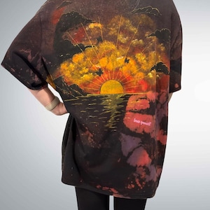 Könnte beinhalten: Ein schwarzes T-Shirt mit kurzen Ärmeln und einer Grafik eines Sonnenuntergangs über dem Wasser mit Silhouetten von Wolken und Schmetterlingen. Das Design zeigt eine gelbe Sonne, einen orange-roten Himmel und den rosa Text "love yourself".