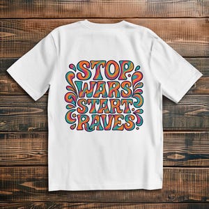Könnte beinhalten: Weißes T-Shirt mit einem farbenfrohen Grafikdesign mit der Aufschrift "STOP WARS START RAVES" in einer Retro-Schriftart. Das Design weist orange, blaugrüne und violette Farbtöne mit dekorativen Wirbeln auf. Das Shirt ist aus weichem Material.