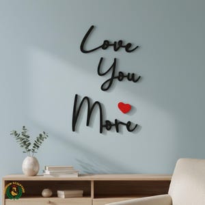 May include: Black cursive wall art that reads "Love You More" with a small red heart. The wall art is mounted on a light blue wall above a wooden cabinet with a vase and books.