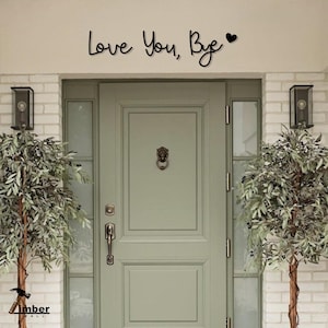 Pode incluir: Uma porta da frente verde sálvia com uma aldrava em forma de cabeça de leão e uma maçaneta de metal preta. As palavras "Love You, Bye" estão acima da porta em fonte cursiva, com um símbolo de coração. Duas oliveiras ladeiam a porta.