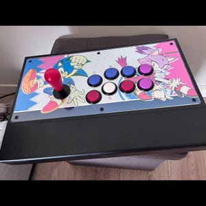 Puede incluir: Un mando arcade negro con una placa gráfica personalizada que muestra a Sonic the Hedgehog y Blaze the Cat. El dispositivo incluye un joystick rojo y ocho botones circulares en azul, blanco, morado y rosa sobre un fondo de rayas blancas.