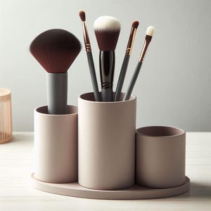 May include: A blush-colored makeup brush holder with three cylindrical compartments and a matching oval base. The holder contains various makeup brushes with dark brown and white bristles, and gray and gold handles.
