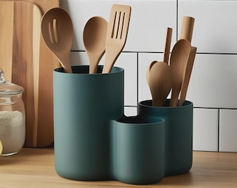 Può includere: Un portautensili verde acqua a tre scomparti, pieno di cucchiai di legno, spatole e altri utensili da cucina. Il supporto è su una superficie di legno, con un tagliere e un barattolo di farina sullo sfondo.