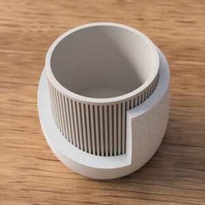 May include: A light gray, modern planter with a cylindrical interior and a textured exterior. The planter features vertical lines around the middle section and sits on a wooden surface. Ideal for small plants.